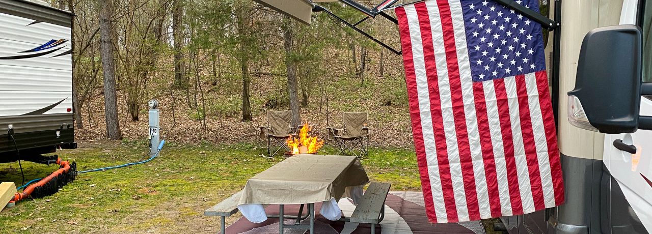 An American flag flies from the side of a RV at a wooded campsite during the day, with a campfire glowing in the background.