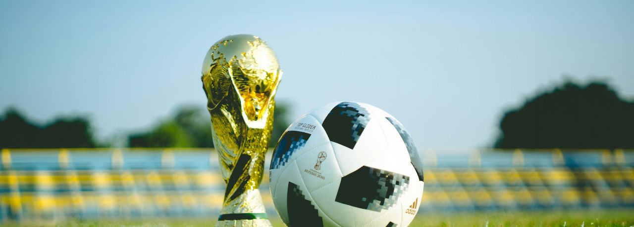 A soccer ball with FIFA World Cup trophy placed on a grassy field with empty stadium seats and trees in the background.