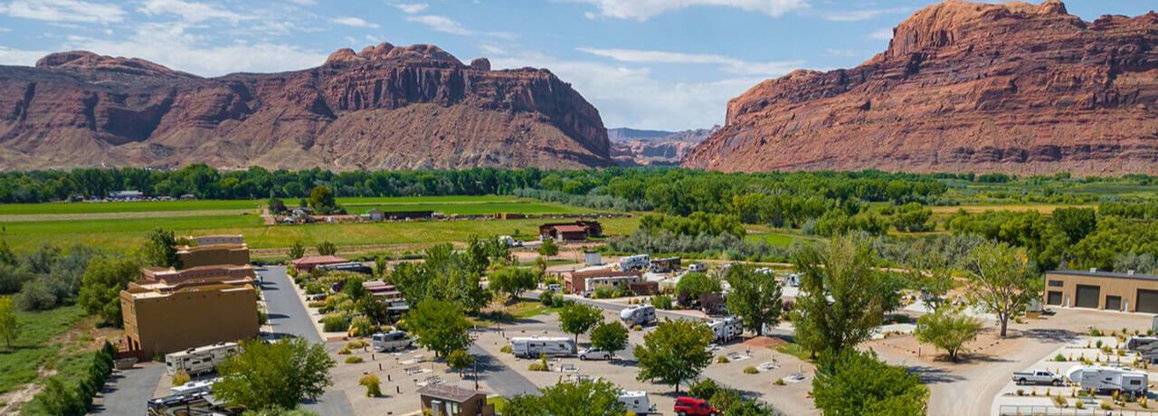 Aerial view of Village Camp Moab with RV sites surrounded by greenery and red rock formations, with the La Sal Mountains visible in the background.