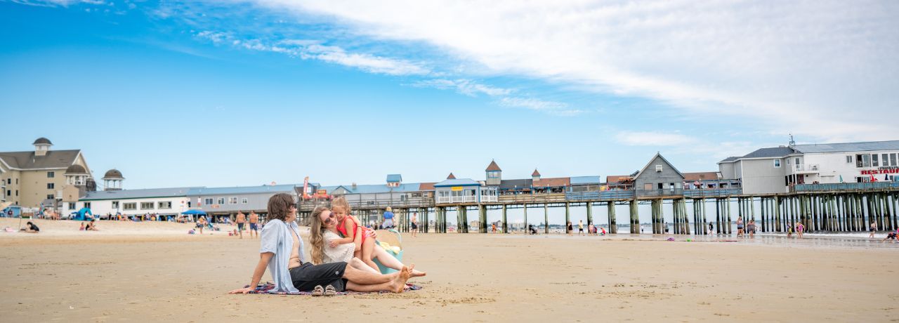Family of 3 with a small child sit on a blanket on a sandy beach near Old Orchard Beach Pier with buildings, under a blue sky with scattered clouds.
